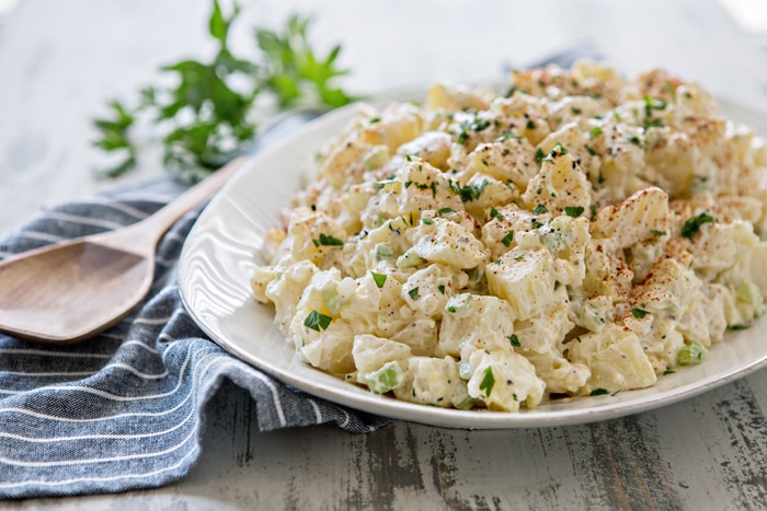 classic potato salad on large white platter next to wooden spoon and blue towel