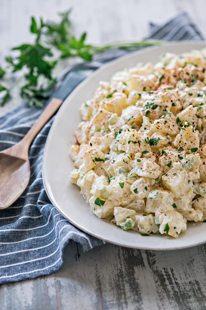 close up shot of classic potato salad on large white platter next to wooden spoon and blue towel