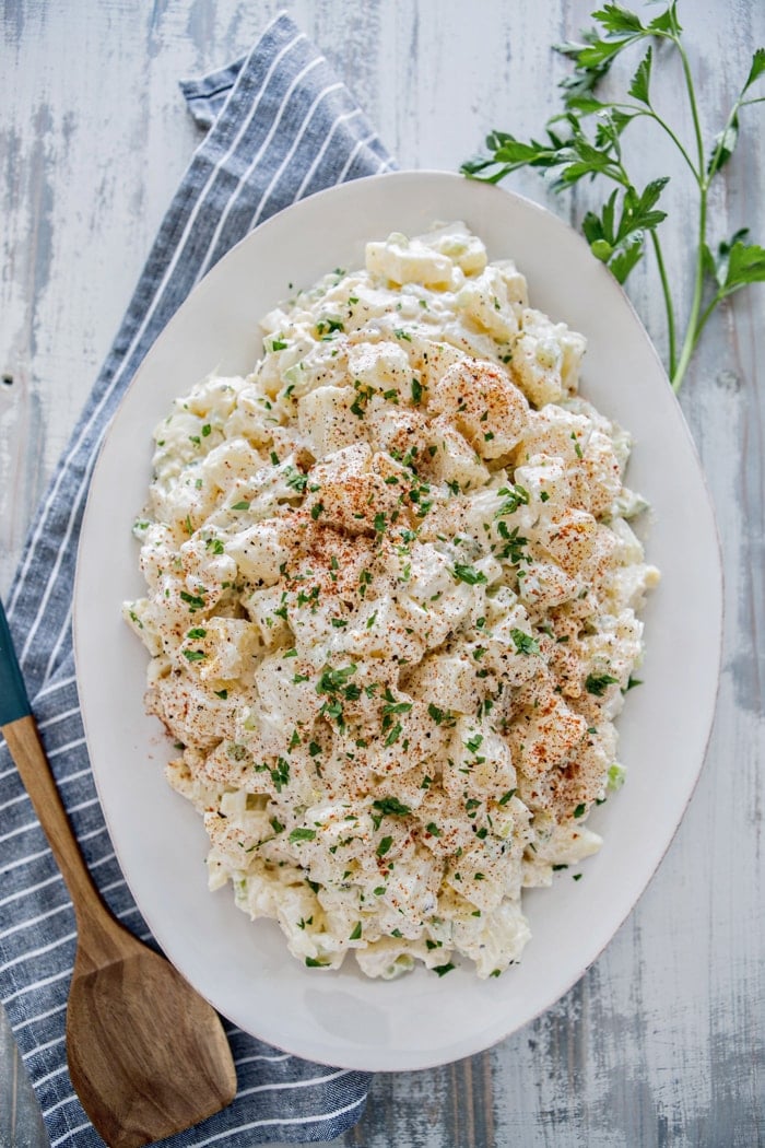 classic potato salad on large white platter sprinkled with parsley next to wooden spoon and blue towel