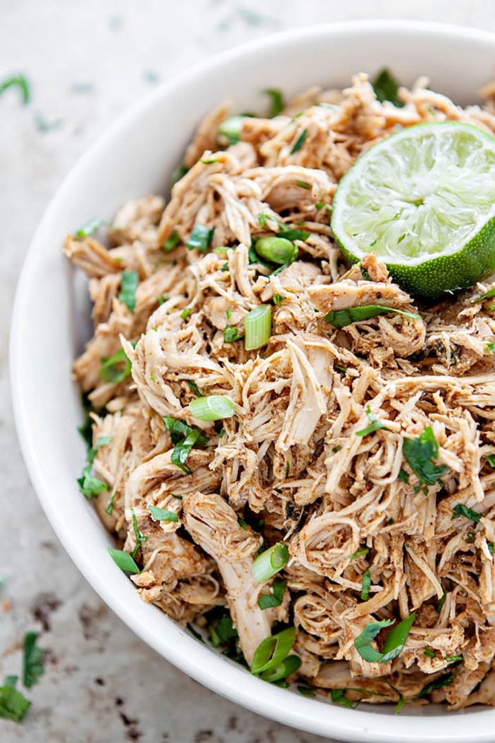 Close-up of shredded Tex-Mex chicken in a white bowl, garnished with chopped green onions, cilantro, and a halved lime. This seasoned chicken is used for homemade chicken enchiladas.