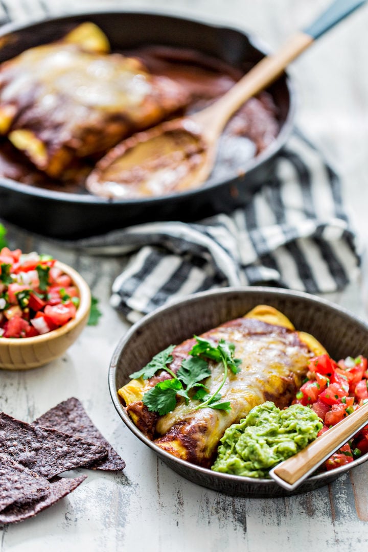 Black skillet with baked homemade chicken enchiladas topped with melted cheese and brown chili gravy. A wooden spoon rests in the pan. A serving of enchiladas is plated with guacamole, pico de gallo, and tortilla chips.