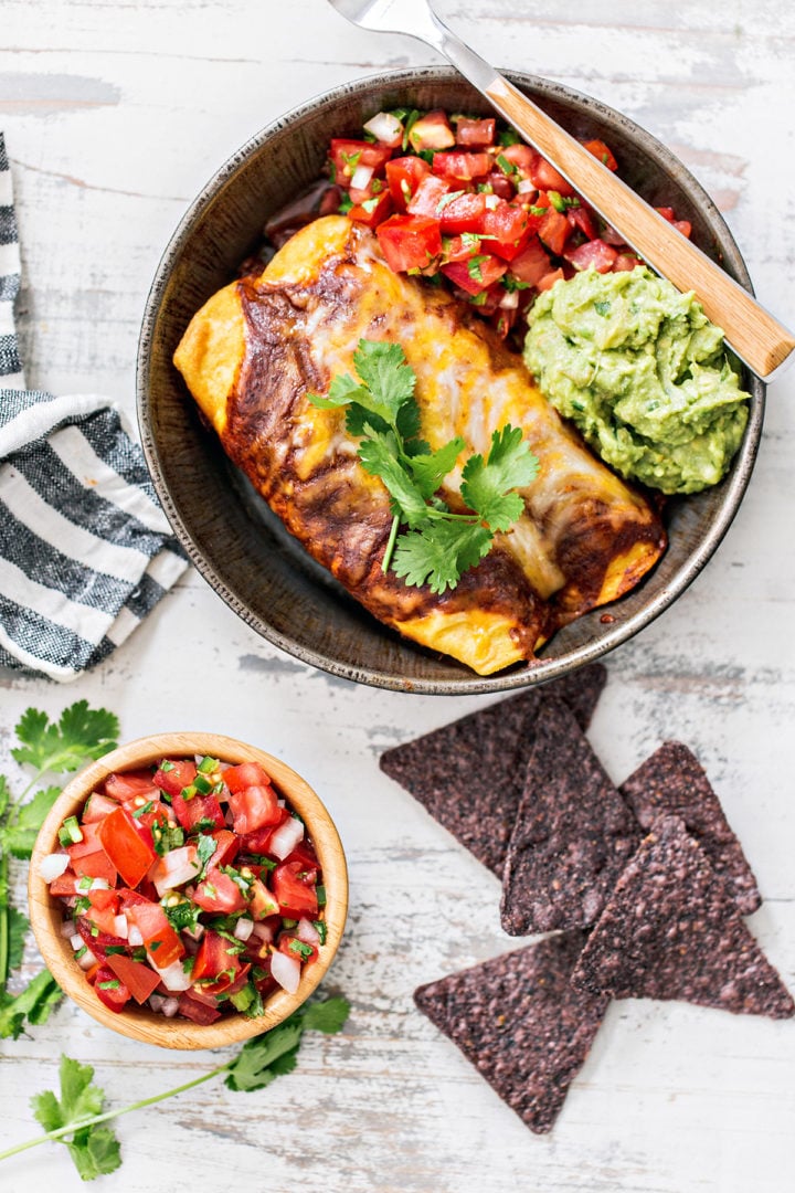 Top-down view of cheesy chicken enchiladas with chili gravy served in a metal dish. Plated with a scoop of guacamole, fresh pico de gallo, and a sprig of cilantro. Blue corn chips and a bowl of pico de gallo sit nearby.