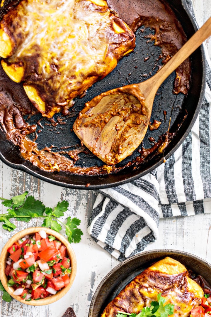 Close-up of a cast iron skillet with partially eaten chicken enchiladas. The melted cheese and rich chili gravy cling to the pan and wooden spoon. A striped towel and bowl of pico de gallo are in the background.