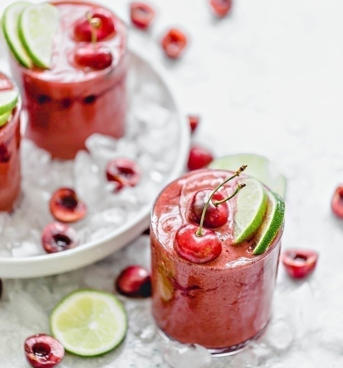 Cherry Limeade Slush in a clear glass surrounded by ice with fresh limes and cherries