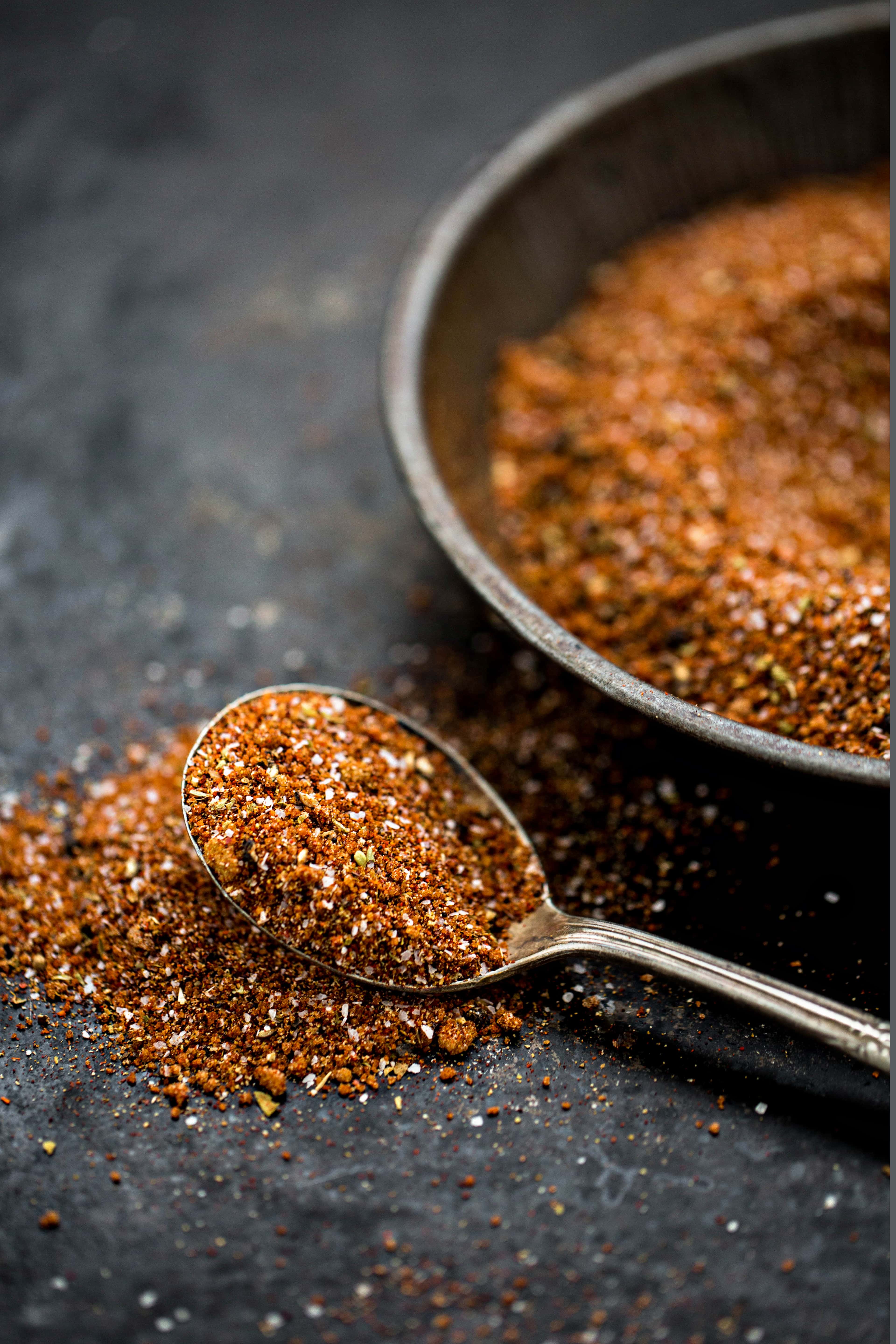 photo of a spoon full of spice rub for smoked pulled pork with a bowl of dry rub in the background on a dark background 