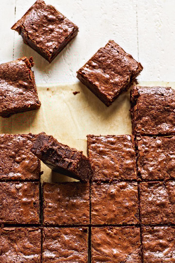 Overhead shot of cut squares of fudgy homemade brownies arranged on brown parchment paper on a white wooden table. One brownie with a bite taken out of it is placed on its side among the other brownies.