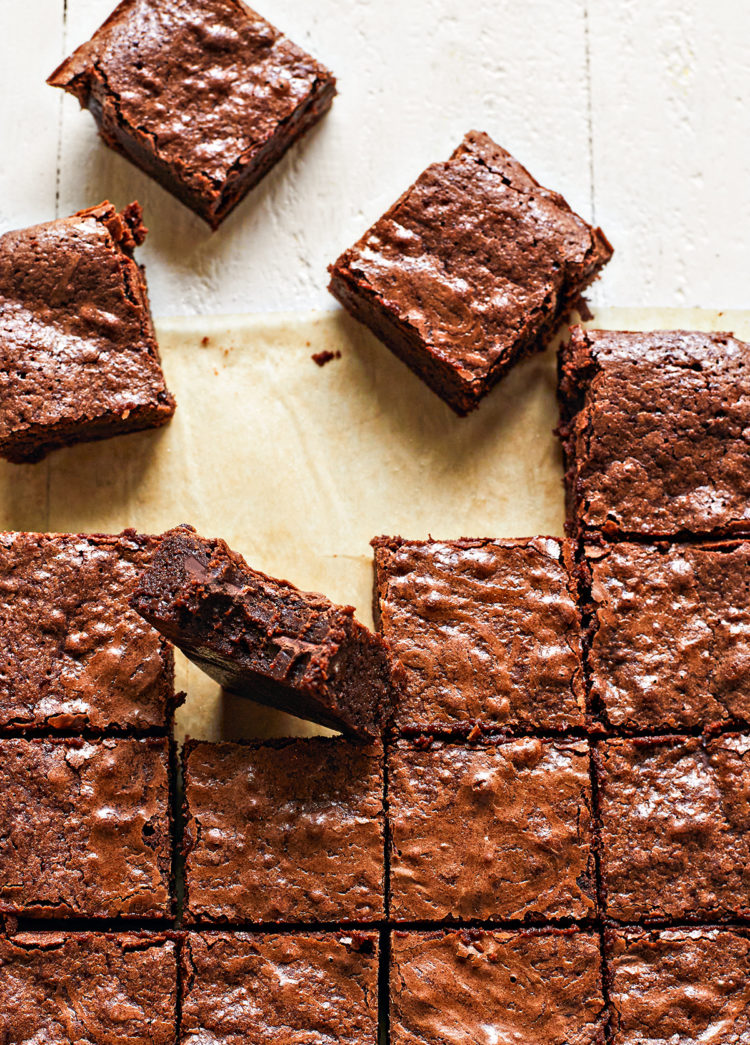 Overhead shot of cut squares of fudgy homemade brownies arranged on brown parchment paper on a white wooden table. One brownie with a bite taken out of it is placed on its side among the other brownies