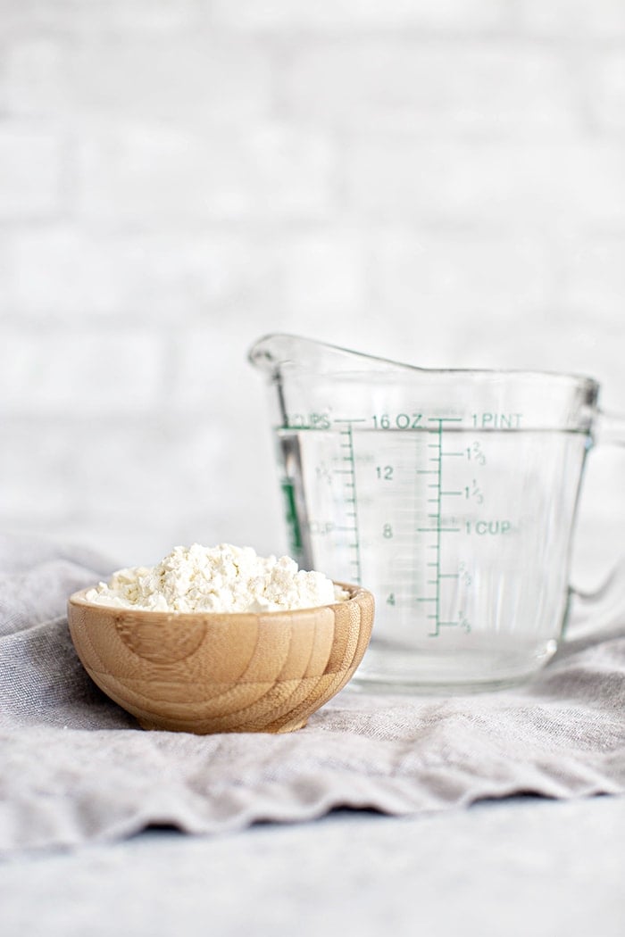 A small wooden bowl filled with buttermilk powder next to a glass measuring cup of water, a convenient shelf-stable alternative to fresh buttermilk.