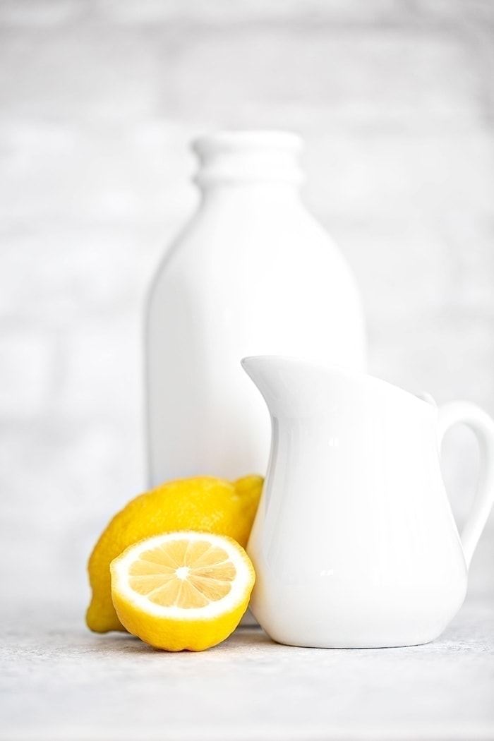 A halved lemon next to a small pitcher of milk, with a milk jug in the background—an easy homemade buttermilk substitute.