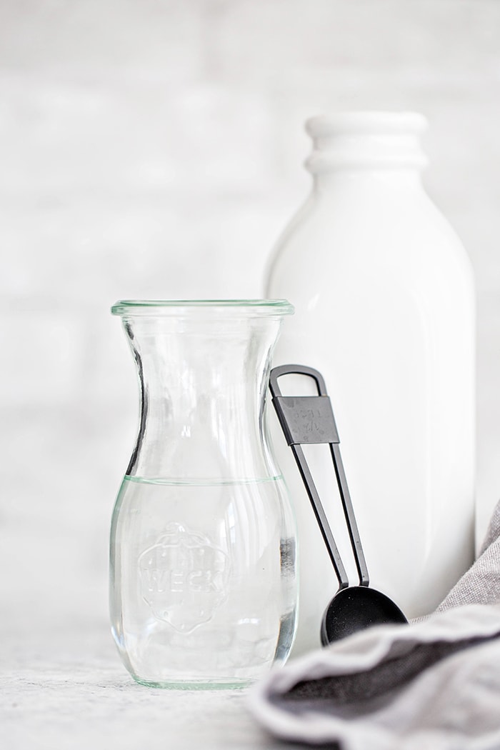 A jar of white vinegar next to a pitcher of milk and a measuring spoon, showing how to make a simple buttermilk substitute.