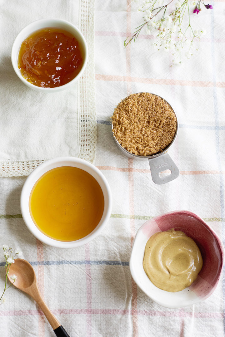 Four small bowls on a pastel plaid tablecloth containing the glaze ingredients: orange marmalade, honey, brown sugar, and mustard. A wooden spoon rests nearby.
