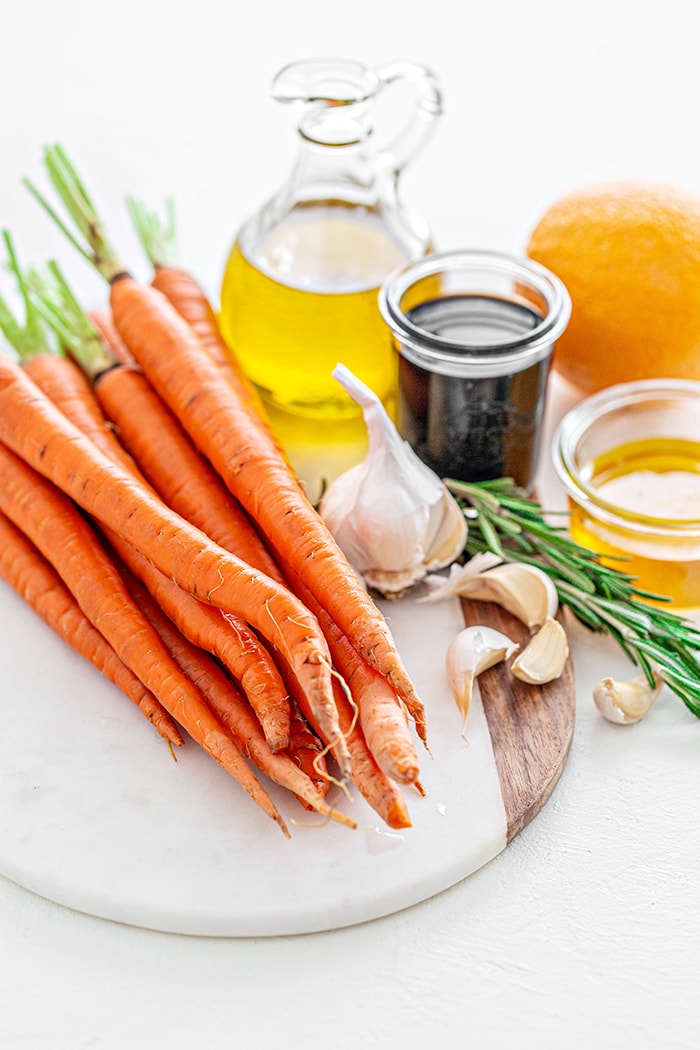 ingredients needed to make Honey balsamic roasted carrots: fresh, whole carrots, garlic cloves, olive oil, honey, orange juice, and fresh rosemary arranged on a marble cutting board