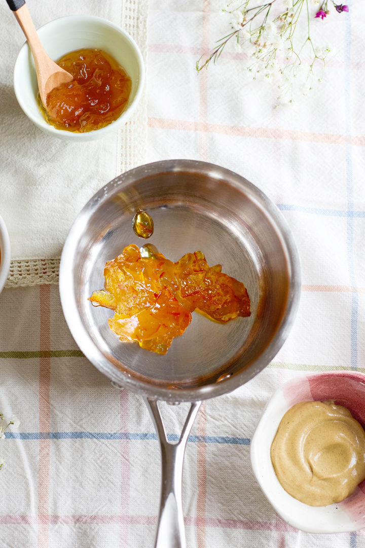 Close-up of a saucepan with orange marmalade inside, ready to be heated. A bowl of coarse mustard sits nearby, partially in frame.