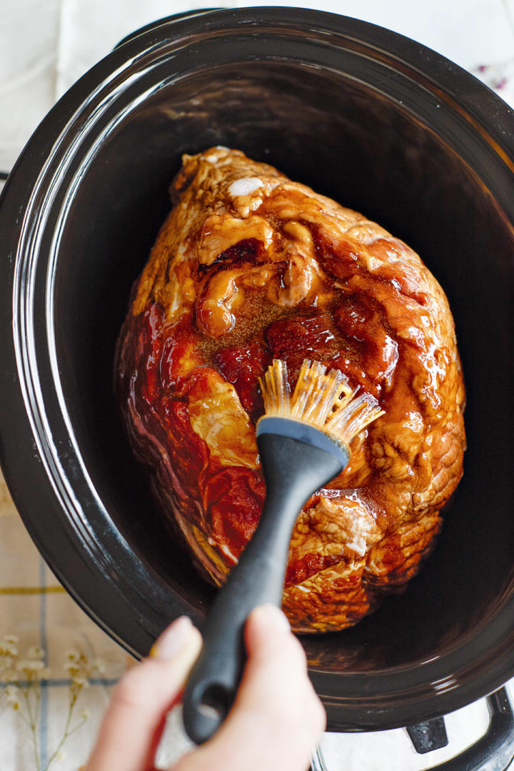 Close-up of a spiral ham in a black slow cooker, with a silicone brush applying the honey mustard glaze over the surface before cooking. The glaze is thick and glossy.