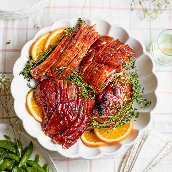 Top-down view of sliced, fully cooked spiral ham arranged in a scalloped white serving dish with orange slices and fresh herbs. The slices are separated and draped outward for presentation.