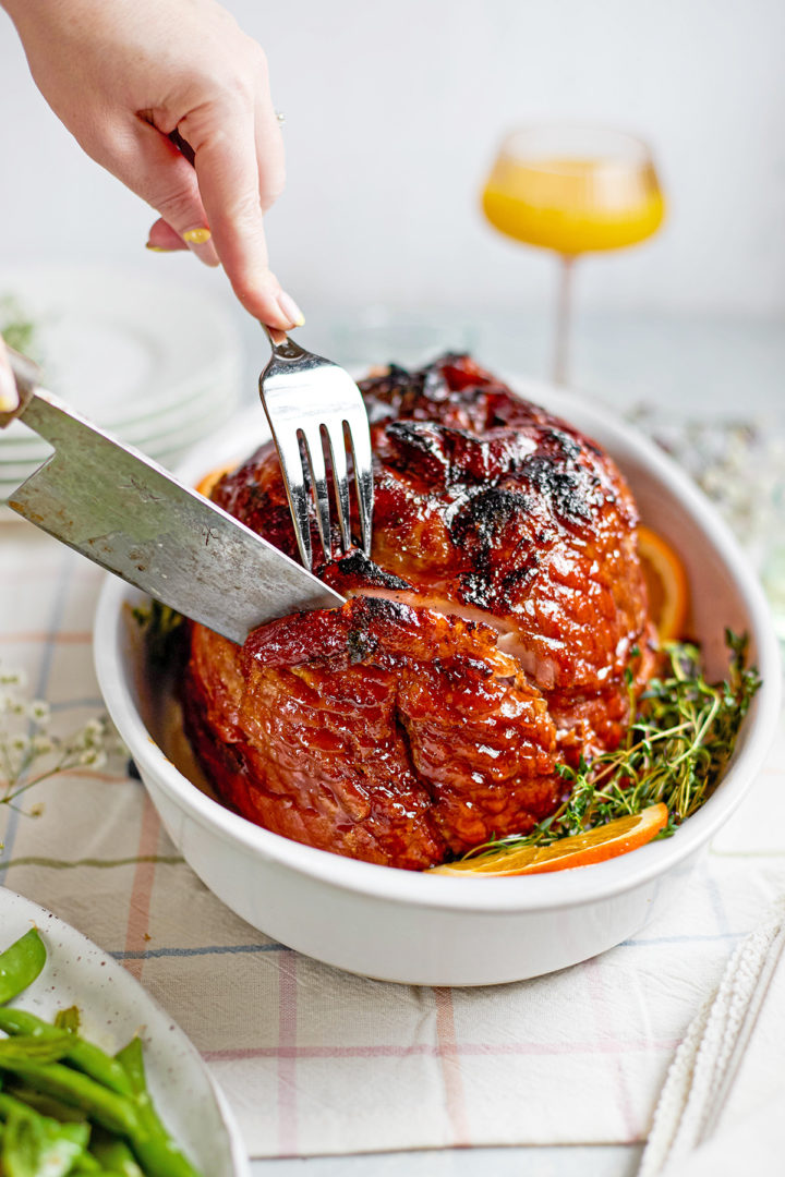 Close-up of a fork and knife carving into the surface of a glazed ham in a white serving dish, showing the deep caramelization on the exterior. This is a slow cooker spiral ham, just before serving.