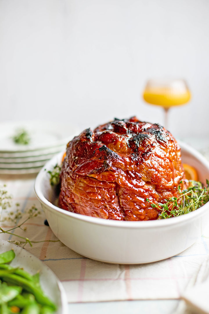 Glazed crockpot spiral ham positioned upright in a white oval baking dish, garnished with fresh thyme and orange slices. The surface is lacquered and slightly charred from broiling.