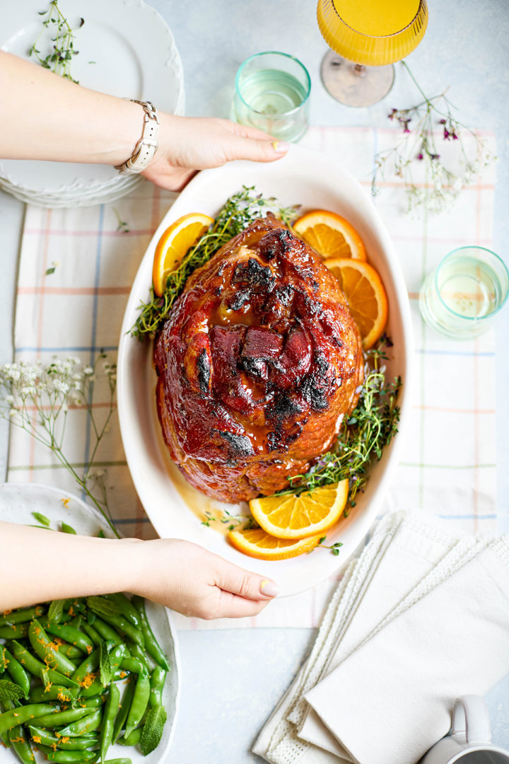 Overhead view of a fully glazed slow cooker spiral ham in an oval white serving dish, surrounded by fresh thyme sprigs and orange slices, with hands setting it on a plaid tablecloth for serving.