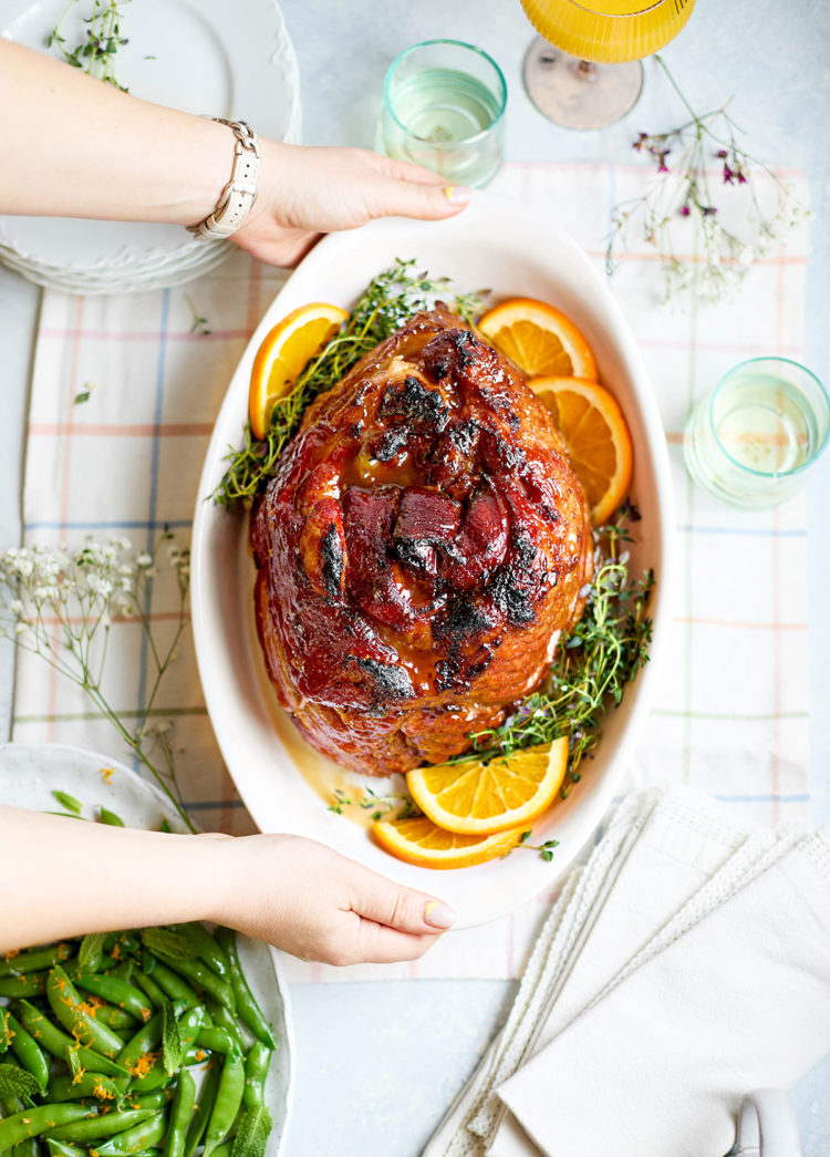 Overhead view of a fully glazed slow cooker spiral ham in an oval white serving dish, surrounded by fresh thyme sprigs and orange slices, with hands setting it on a plaid tablecloth for serving.