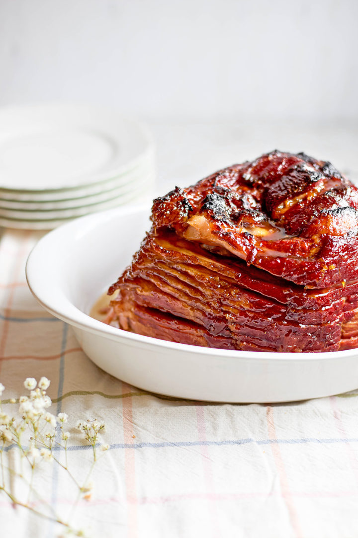 Slow cooker spiral ham resting in a white baking dish, glistening with glaze and caramelized edges, showcasing the layers of the ham as they fan out horizontally. A stack of white dinner plates is blurred in the background.