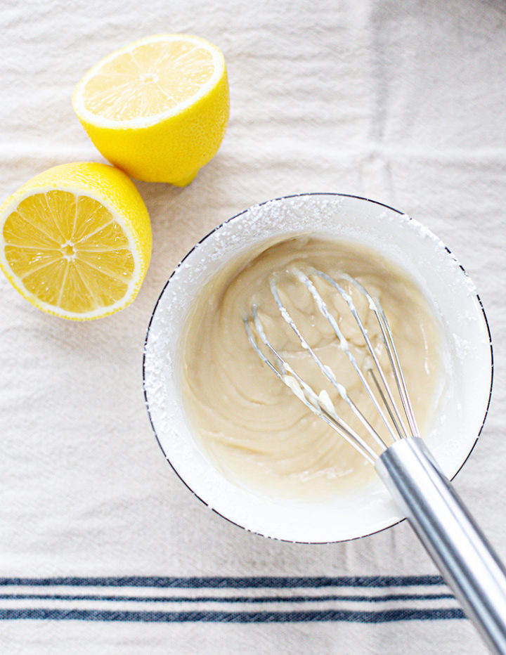 Whisking Lemon Glaze in Bowl: A metal whisk rests in a bowl of smooth lemon glaze, freshly mixed for drizzling over blueberry lemon scones. Two halved lemons sit beside the bowl on a striped linen towel, highlighting the fresh citrus flavor used in the glaze.