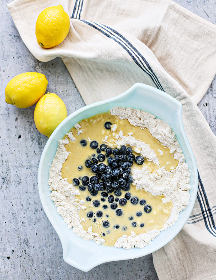 Wet and Dry Ingredients in Mixing Bowl: A pale blue mixing bowl filled with flour, wet ingredients, and a pile of fresh blueberries before combining—one of the first steps in how to make blueberry lemon scones from scratch. Three whole lemons rest on a nearby kitchen towel, setting a bright, citrusy scene.
