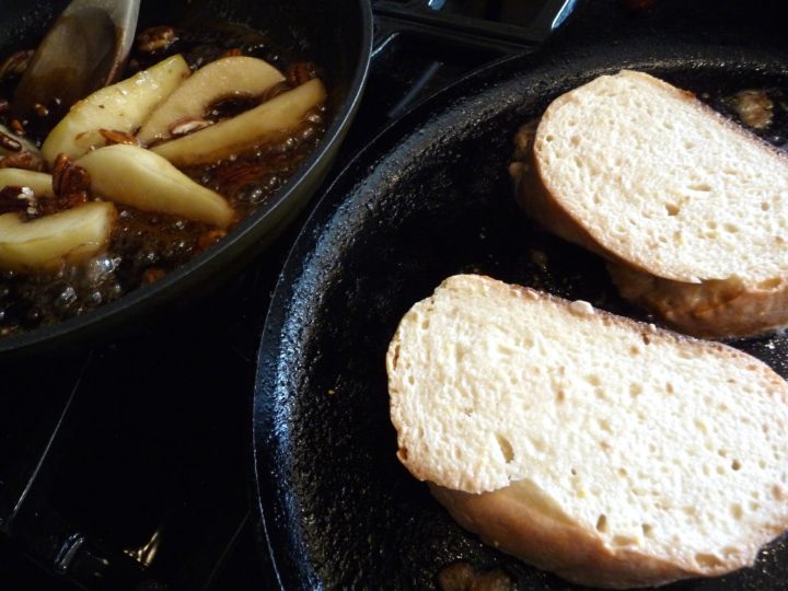 Two pieces of french toast being cooked in a skillet. Another skillet contains pears and pecans.