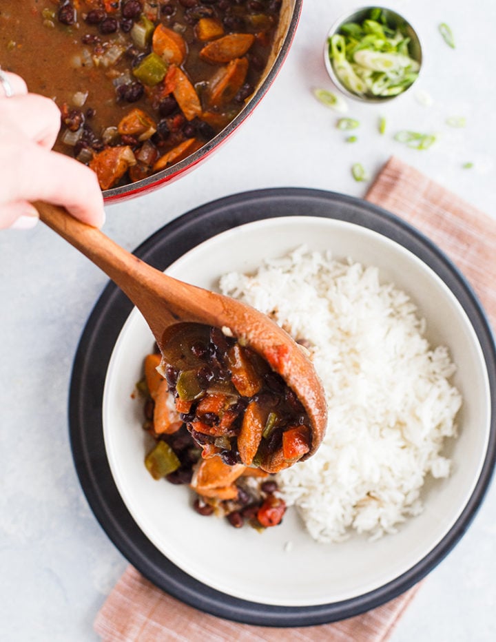 woman using wooden spoon to serve black beans and smoked sausage on to a bowl of white rice