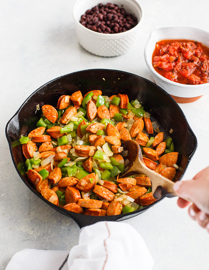 woman stirring sausage, peppers, and onions in a cast iron pan for a black beans and rice recipe