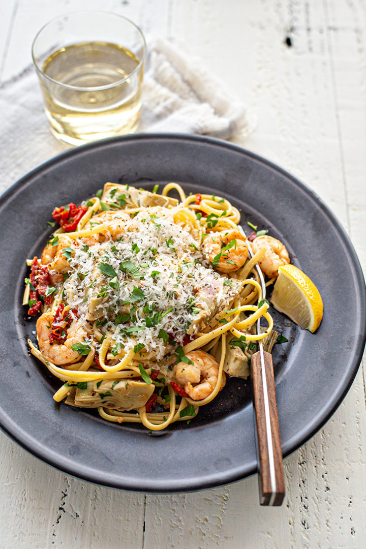 plate with shrimp scampi with linguine and artichokes on a white farmhouse table with a glass of wine