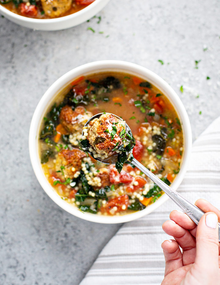 woman holding a spoon with a meatball in it over a white bowl with italian wedding soup  