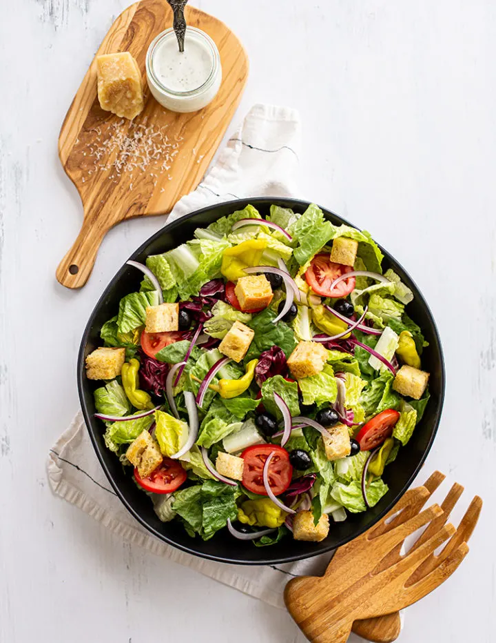 overhead shot of a bowl of olive garden salad recipe with a jar of olive garden dressing on a cutting board with parmesan cheese