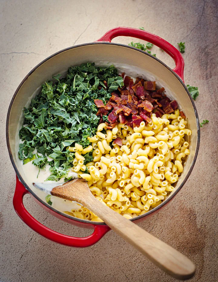 a pot with macaroni noodles, bacon, kale, and white cheddar cheese sauce for a recipe for white cheddar mac and cheese