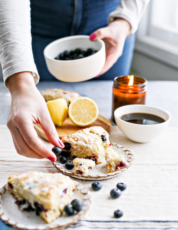Photo of hand reaching for a blueberry near plated scone: A close-up of a hand placing fresh blueberries next to a glazed lemon blueberry scones on a rustic ceramic plate. A candle, sliced lemon, and a bowl of blueberries are in the background, creating a cozy breakfast scene.