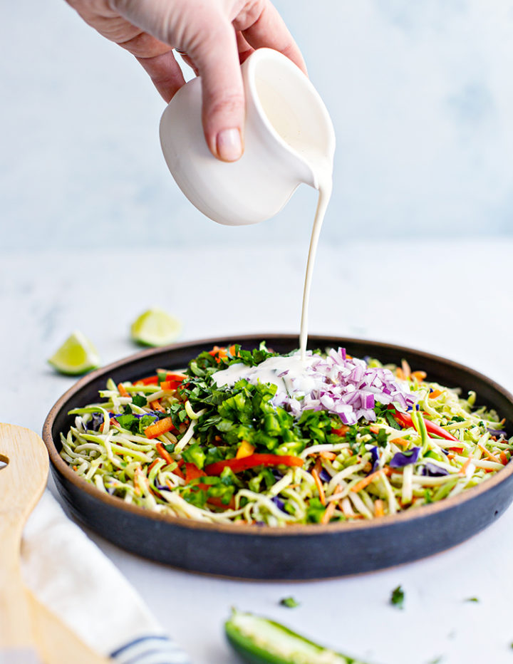 photo of woman's hand pouring creamy cilantro lime dressing on top of a bowl of broccoli slaw