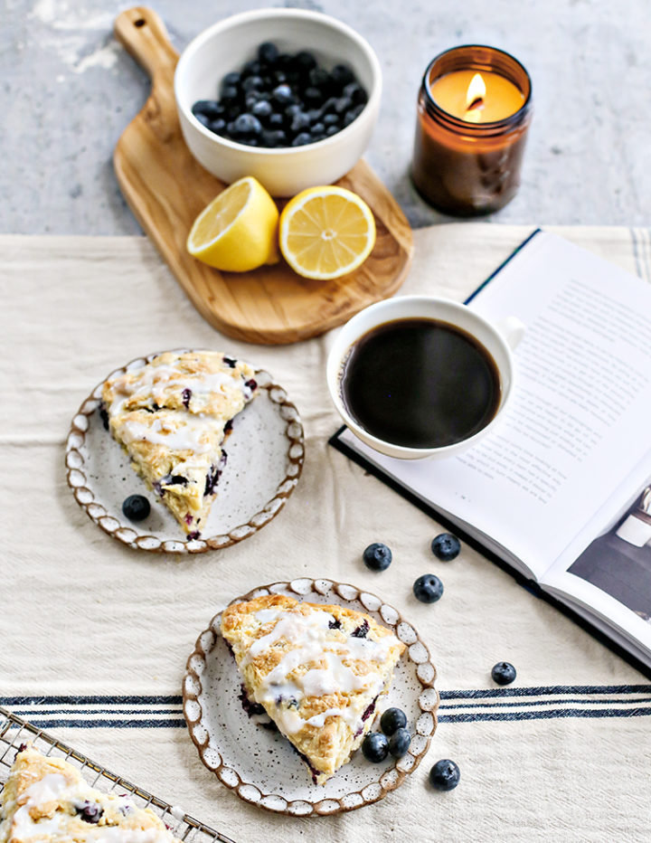 Flatlay of plated scones with coffee and book: Overhead shot of blueberry lemon scones with lemon glaze served on small ceramic plates beside a hot cup of black coffee, fresh blueberries, a halved lemon, and a lit candle. A book is open beside the plate for a relaxed brunch mood.