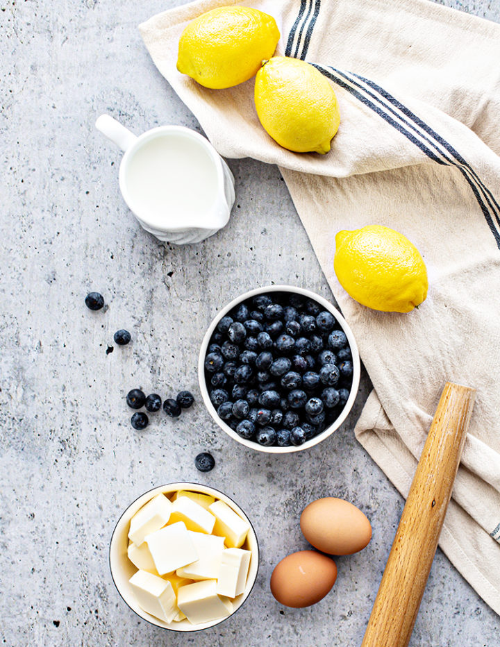 Flatlay photo showing the ingredients for blueberry lemon scones: cubes of cold butter, fresh blueberries, whole lemons, eggs, and milk arranged on a textured surface with a rolling pin and striped linen.