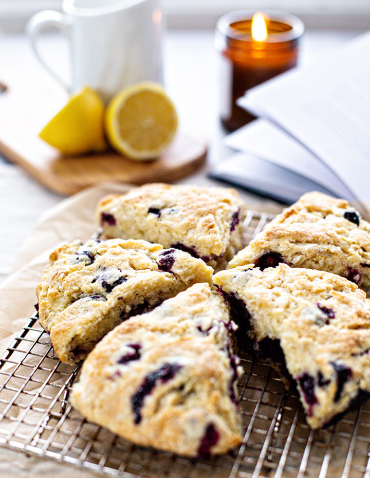 Baked scones cooling on rack with lemons and coffee in background: Golden-brown lemon blueberry scones cooling on a wire rack. A mug of coffee, halved lemon, and lit candle create a warm, inviting kitchen scene.