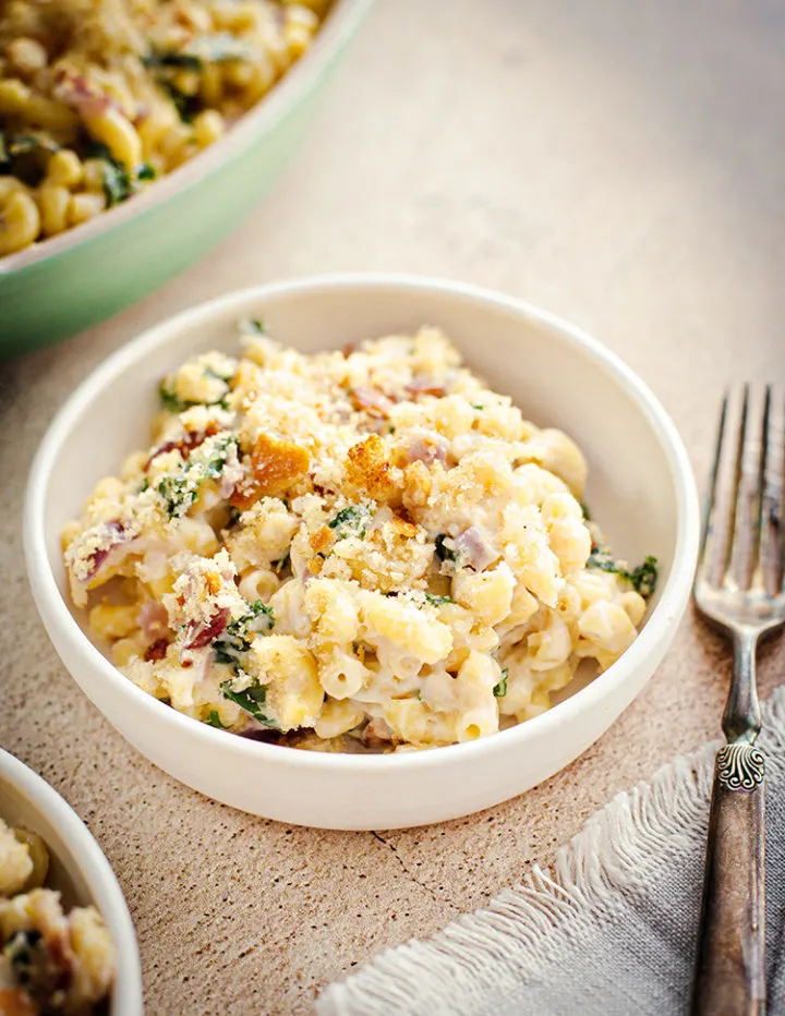 a close up shot of a white bowl with White Cheddar Mac and Cheese with kale and bacon