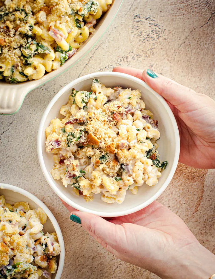 an overhead shot of a woman holding a white bowl with White Cheddar Mac and Cheese with kale and bacon