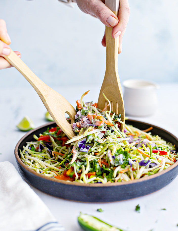 photo of a woman's hands stirring a bowl of cilantro lime broccoli slaw with wooden spoons