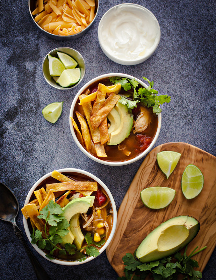 overhead shot of styled bowls of chicken tortilla soup on a counter with limes, cheese, and sour cream
