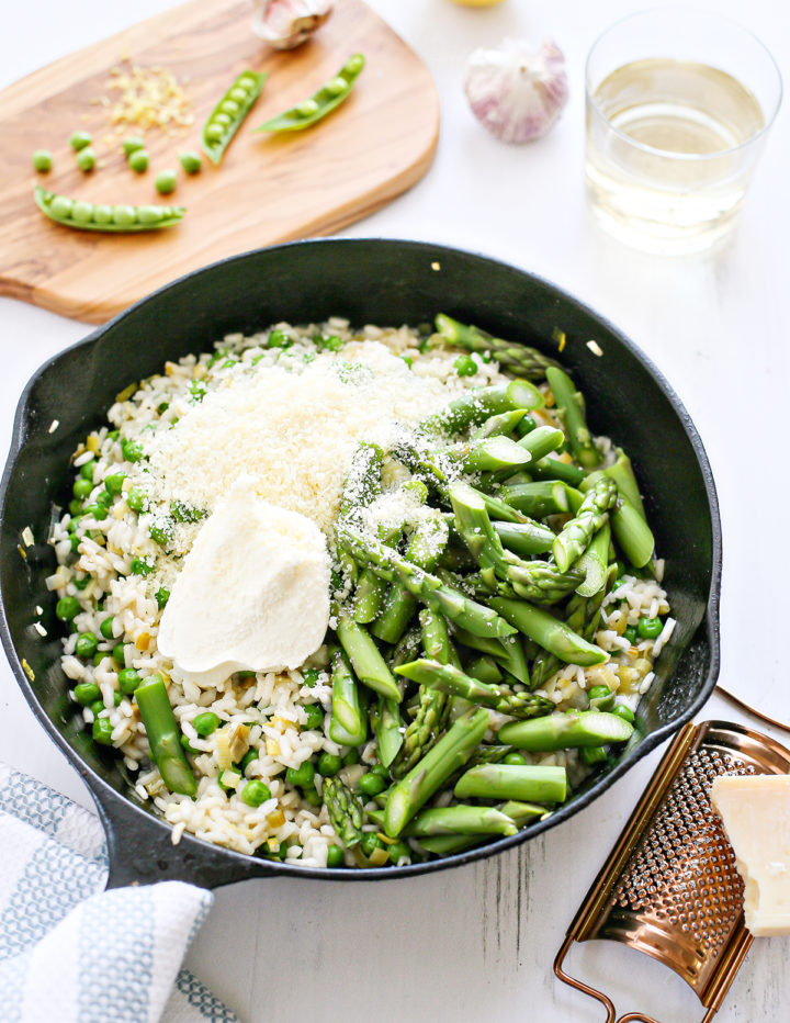 Cast iron pan filled with asparagus risotto just before stirring in the final ingredients. Topped with a generous scoop of mascarpone, grated parmesan, and tender asparagus and peas. Garlic, lemon, and wine in the background set the scene for a fresh spring dinner.