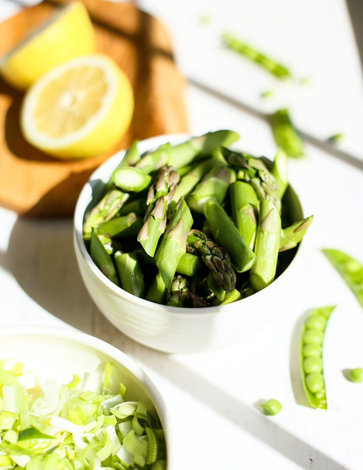 A white bowl filled with freshly chopped green asparagus, ready for cooking. The background includes bright lemon halves on a wooden board and a scattering of sliced sugar snap peas, setting a fresh springtime mood for this lemon asparagus risotto recipe.