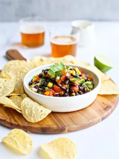 photo of black bean salad in a bowl on a cutting board surrounded by tortilla chips and glasses of beer