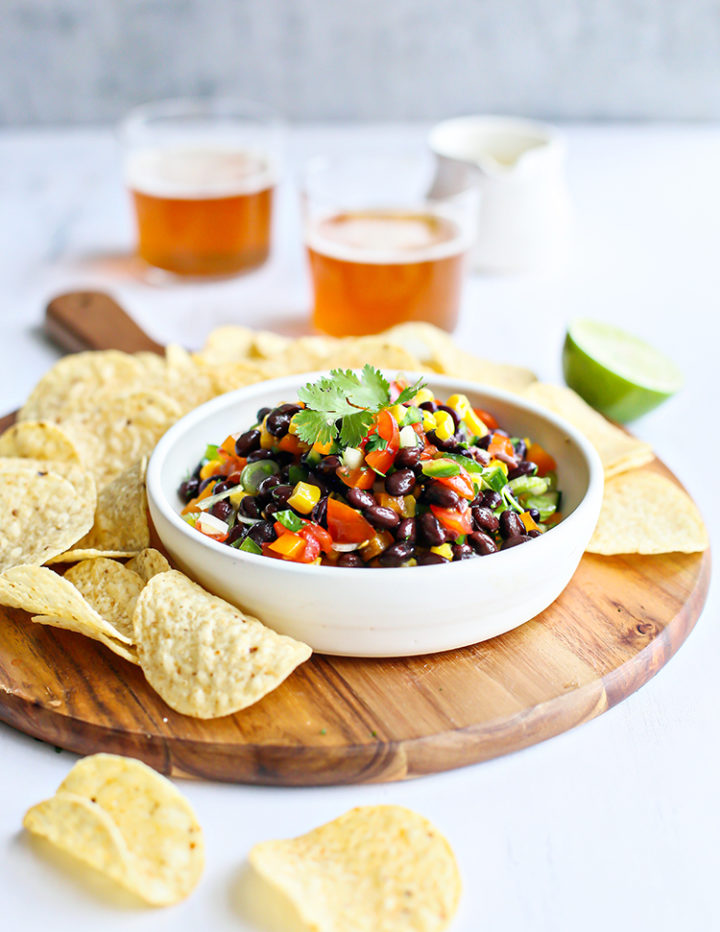photo of black bean salad in a white bowl on a cutting board surrounded by tortilla chips and glasses of beer