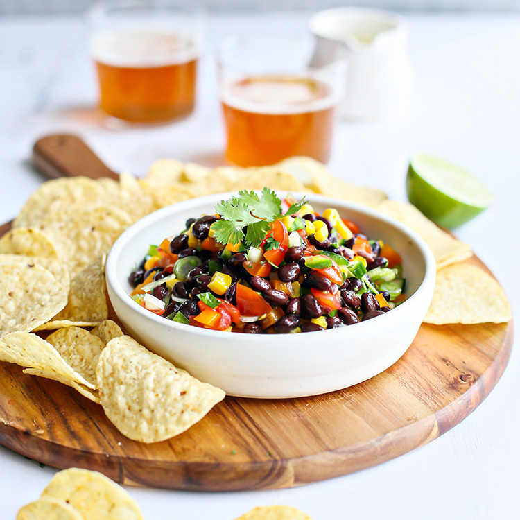 photo of black bean salad in a bowl on a cutting board surrounded by tortilla chips and glasses of beer