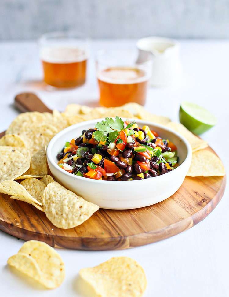 photo of black bean salad in a bowl on a cutting board surrounded by tortilla chips and glasses of beer