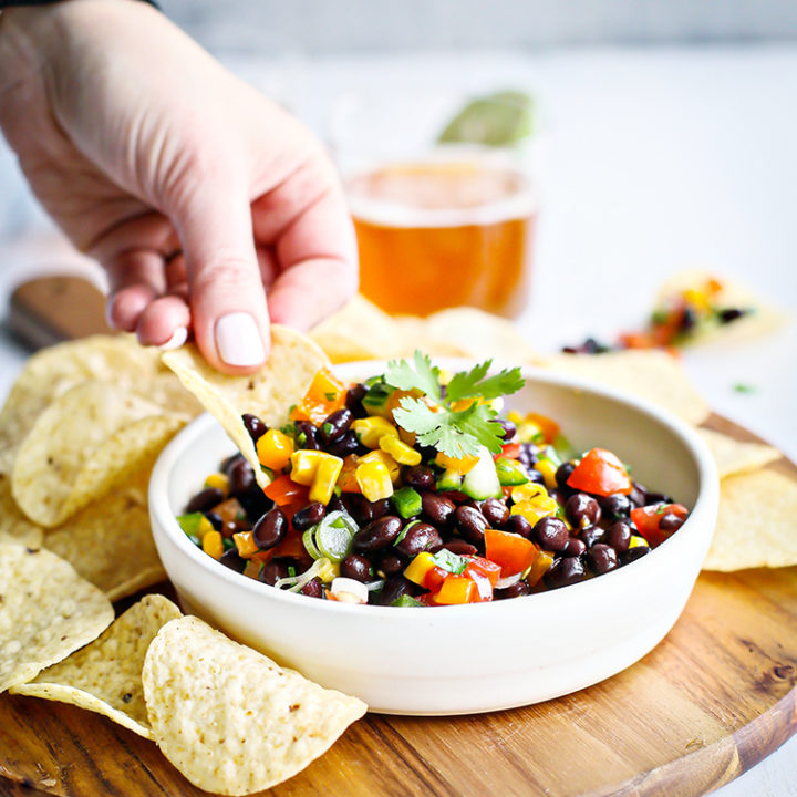 photo of black bean salad in a white bowl with woman scooping with a tortilla chip