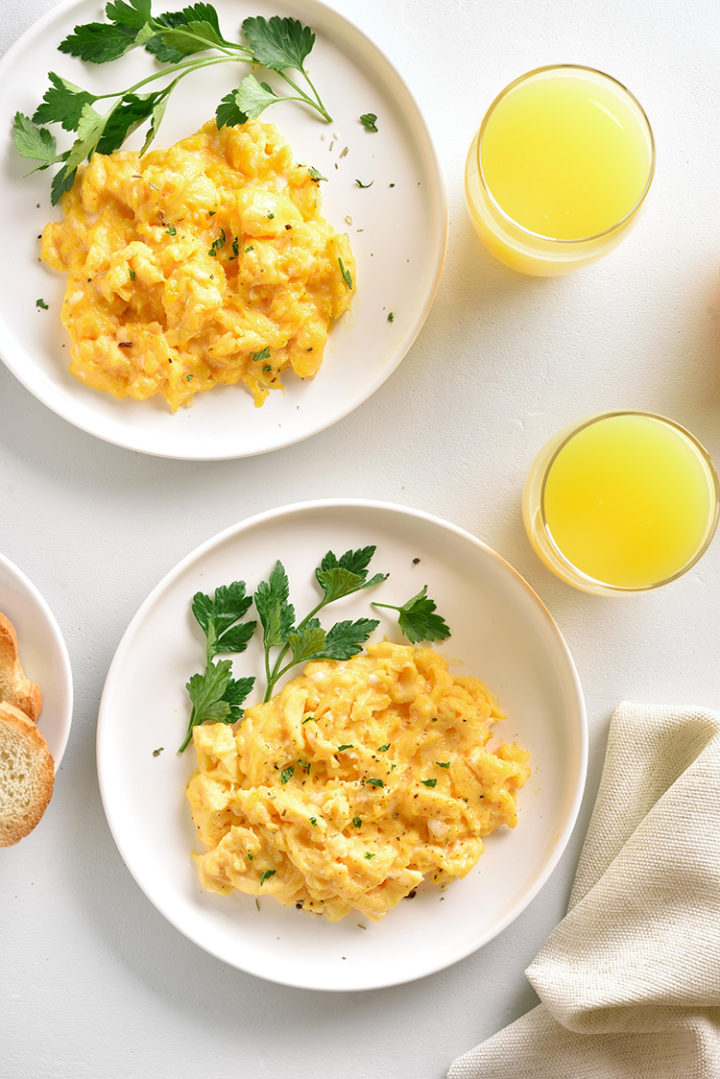 an overhead shot of two white plates with scrambled eggs and parsley on a kitchen table with two glasses of orange juice